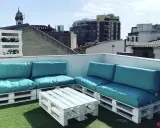Sofá y mesa de centro hechos con palets blancos, con cojines turquesas, en una terraza urbana. ¡Una idea genial de como hacer un sofa con palets!