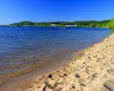 Słoneczny dzień nad zalewem Pilica. Piaszczysta plaża, spokojna woda i zielone wzgórza w tle. Kilka osób pływa i odpoczywa.