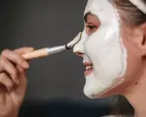Kobieta nakłada białą maskę na twarz pędzelkiem. Składnik cetrimonium chloride w kosmetykach pielęgnacyjnych pomaga w nawilżeniu skóry.