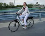 Młoda kobieta w białych spodniach i jasnym swetrze jedzie na rowerze elektrycznym, ciesząc się jazdą i osiągając swoją maksymalną prędkość.