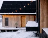 Ett modernt solcellsbatteri utomhus monterat på en träfasad, med snö på taket och marken. Julbelysning lyser upp kvällen.