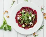Eleganckie carpaccio z buraka z fetą, orzechami i rukolą na białym talerzu, otoczone świeżymi ziołami i orzechami.