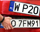 Dłoń z bransoletką montuje tablice rejestracyjne na czerwonym samochodzie. Widać dwie tablice: jedną z napisem WP20, drugą z numerem O-7FM91.