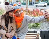 Para do selfie w zimowych stylizacjach. Ona w beżowym płaszczu i czapce z pomponem, on w szarej bluzie i czapce z pomarańczowym akcentem. Jaka czapka do płaszcza? Ta jej wygląda świetnie!