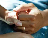 Två händer håller varandra, en gest som visar medlidande och stöd. En tunn ring syns på ena handen.