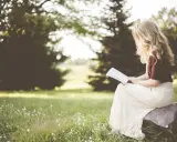 Kobieta z blond włosami czyta psalm 6 tekst, siedząc na kamieniu w trawie, otoczona zielenią.