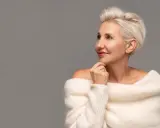 Le visage d'une femme de 80 ans, aux cheveux blonds courts, porte un pull blanc en maille. Elle regarde sur le côté, la main sous le menton.