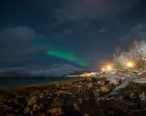 Nocne niebo nad zaśnieżonym wybrzeżem, gdzie zobaczyć zorzę polarną. Zielona poświata tańczy nad górami, a światła latarni oświetlają kamienisty brzeg.
