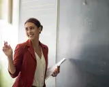 Lehrerin mit Kreide und Unterlagen vor einer Tafel mit einer Matheaufgabe. Sie lächelt und wirkt engagiert, was auf einen interaktiven Unterricht hindeutet.