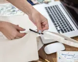 Ręce tworzą torbę DIY, przyszywając czarną wstążkę do płóciennego materiału. Obok leży laptop i myszka.
