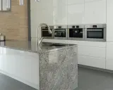 Nowoczesna kuchnia z kamienia. Wyspa kuchenna z granitowym blatem i zlewem, obok zabudowa z piekarnikami i ekspresem do kawy.