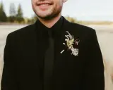 A groom in all black wedding guest attire men's suit, complete with a black tie and a rustic boutonniere.