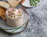 Un pot de rillettes de saumon crémeuses, garni de ciboule, prêt à être tartiné sur des toasts grillés.