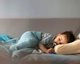 Mały chłopiec śpi na kanapie, otulony kocykiem. Wygląda na to, że mimo początkowego buntu, kiedy dziecko rezygnuje z drzemki, w końcu uległ zmęczeniu.