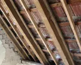Charpente en bois avec des tuiles rouges et un mur en briques décrépit. Les contre latte sont visibles, soutenant la structure.