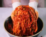 Ręce w białych rękawiczkach trzymają głowę kapusty pekińskiej pokrytą czerwoną pastą. To właśnie kimchi, tradycyjna koreańska potrawa.