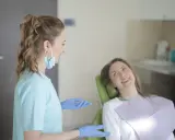Dentystka w niebieskiej bluzie i rękawiczkach rozmawia z pacjentką. Długie studia stomatologiczne wymagają zaangażowania.