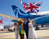 Två kvinnor och en man med en björndräkt firar framför ett flygplan från Norse Atlantic Airways. En kvinna håller en amerikansk flagga, en annan en brittisk flagga.