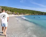 En man går längs en vit sandstrand med turkost vatten. Palmklippor och parasoll syns i bakgrunden. En perfekt plats att undra var ligger Curacao.