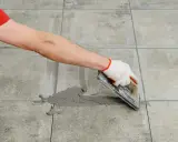 Mano con guante aplica lechada gris a las juntas entre azulejos de cemento con una llana.