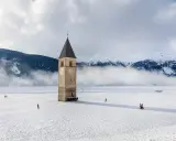 Torre sommersa nel lago di Resia, circondata da neve e montagne nebbiose. Alcune persone passeggiano sul ghiaccio.