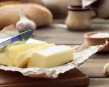 Ein Messer schneidet ein Stück von einem Block welcher Butter. Daneben liegen Knoblauch und ein Brot.