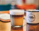 Ein geschichteter Latte Macchiato im Glas, daneben eine weiße Tasse mit Kaffee-Aufdruck.
