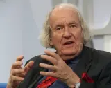 Ein älterer Mann mit langen, hellen Haaren spricht gestikulierend in einem Anzug.