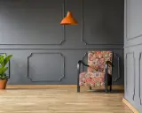 Nowoczesne sztukateria ścienna inspiracje: fotel w kolorowe wzory, pomarańczowa lampa i roślina na tle szarej ściany.