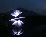 Un artista crea forme luminose con la light painting fotografia, disegnando spirali e petali di luce viola su uno sfondo scuro con montagne e riflessi sull'acqua.