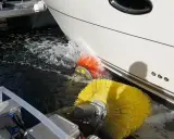 Nettoyage de la partie immergée d'un bateau avec des brosses rotatives jaunes et oranges. L'eau éclabousse autour des brosses.