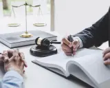 Dłoń prawnika pisząca w księdze, obok wagi sprawiedliwości i młotka sędziowskiego.