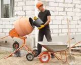 Budowlaniec wsypuje składniki do betoniarki, przygotowując beton B30. W tle taczka i łopata.
