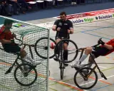 Gracze w piłkę rowerową walczą o piłkę przy bramce. Jeden z zawodników próbuje strzelić gola.