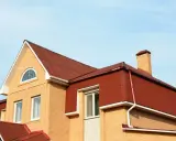 Dach mansardowy konstrukcja z czerwonej blachy falistej, z ceglaną ścianą i kominem.