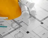 Żółty kask budowlany, zwinięte plany i przybory kreślarskie na projekcie domu. Może posłużyć jako wzór zgłoszenia samowoli budowlanej.