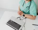 Lekarka w zielonym fartuchu z latarką i laptopem przed sobą. Czy do ginekologa potrzebne jest skierowanie? Zapytaj online.