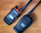 Dwa radiotelefony, jeden pomarańczowo-czarny RETCIS, drugi czarny Midland, leżą na drewnianym blacie.