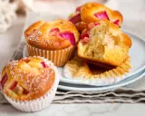Saftige Rhabarber Muffins, mit Puderzucker bestäubt, liegen auf einem Teller. Einer ist angeschnitten und zeigt die rosa Rhabarberstücke.