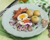 En tallrik med sill, ägg, prinskorv, potatis och rom. En härlig påskbord mat som ser utsökt ut.