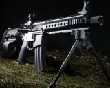 Czarny karabin AR z magazynkiem bębnowym i dwójnogiem, leżący na zielonej siatce maskującej.