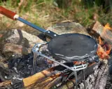 En gjutjärnspanna med trähandtag står på en ställning över en öppen eld. Perfekt för att steka plättar eller våfflor ute i naturen.