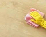 En hand i gul handske torkar av ett ljust trägolv med en rosa trasa. Perfekt för pigmenterad olja till ekbord.