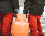 Dwóch mężczyzn w pomarańczowych spodniach niesie butlę z gazem. Wygląda na to, że nie można zakręcić butli z gazem, co może być problemem.