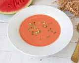 Un refrescante gazpacho de sandía, adornado con trocitos verdes, listo para disfrutar junto a una rodaja de sandía y una cuchara dorada.