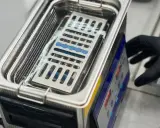 Ein Handschuh bedient ein Gerät, das zeigt, wie funktioniert ein ultraschallreiniger. Kleine Instrumente liegen in einem Korb im Wasserbad.