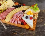 Apetyczna deska serów i wędlin: salami, szynka, ser żółty, pleśniowy, oliwki, winogrona, maliny, orzechy i krakersy.