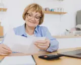 Uśmiechnięta starsza pani z okularami przegląda dokumenty, planując swoje przejście na emeryturę. Obok leży kalkulator i laptop.