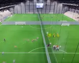 Barn tränar fotboll på en grön konstgräsplan i Prioritet Serneke Arena. Publik sitter på läktaren.