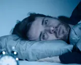 Mann liegt wach im Bett, Augen weit offen, die Angst vor Schlaflosigkeit ist ihm ins Gesicht geschrieben. Ein Wecker ist im Vordergrund zu sehen.